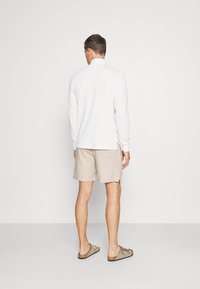 White long-sleeve shirt with side slits, paired with beige shorts. Individual wears brown slide sandals, viewed from the back against a plain background.