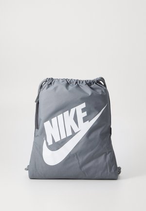 Sac à cordon Nike gris avec grand logo blanc sur le devant et poche zippée latérale, posé sur un fond clair uni.
