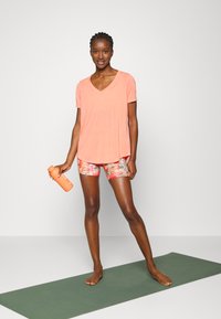 T-shirt coral de decote em V, em tecido leve, combinado com calções de padrão floral. O modelo segura uma garrafa de água laranja, de pé sobre um tapete verde.