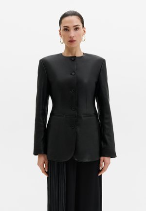 Femme aux cheveux foncés tirés en arrière porte une veste en cuir noire boutonnée avec des épaules structurées et un pantalon noir plissé sur fond blanc.