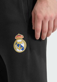 Pantalones deportivos negros con un logo bordado a color del Real Madrid, incluyendo una corona dorada sobre el emblema circular en el muslo.