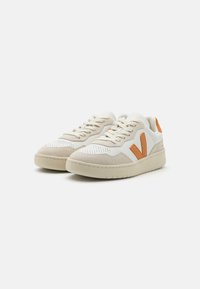 Sneaker beige e bianche con tomaia perforata, accenti arancioni, chiusura con lacci e suola testurizzata, con un grande logo 'V'.