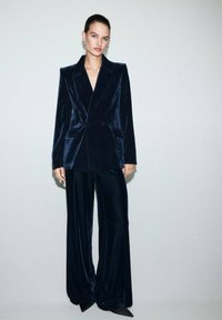 Una mujer se encuentra de pie frente a un fondo liso, vistiendo un traje de terciopelo azul oscuro con un blazer de doble botonadura y pantalones de pierna ancha, combinado con zapatos de punta negra.