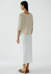 Beiger Strickpullover mit 3/4-Ärmeln, kombiniert mit einem weißen Leinenrock. Verfügt über einen abgerundeten Saum und ein kleines Knopfdetail hinten. Braune Absatzclogs.