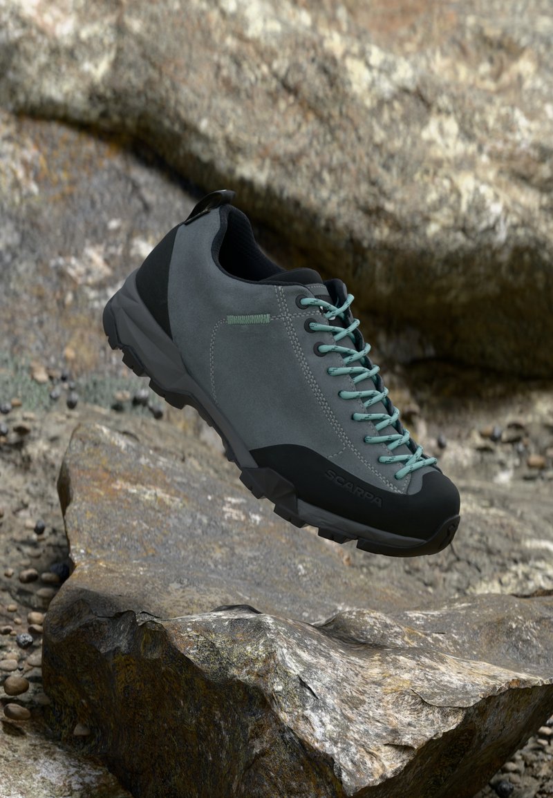 Graue Wildleder-Wanderschuhe mit schwarzen Akzenten, strukturiertem Sohle, türkisfarbenen Schnürsenkeln und verstärkter Zehenpartie. Positioniert über einer steinigen Oberfläche.