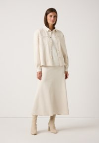 Crème-kleurige button-up overhemdjasje met twee voorzakken, gelaagd over een lichte trui en lange rok, gecombineerd met kniehoge laarzen.
