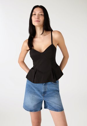 Kobieta ubrana w czarny bezrękawnik z baskinką i niebieskie szorty dżinsowe, stojąca z rękami za plecami na jednolitym tle.