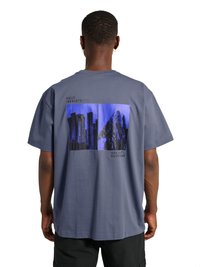 Homme portant un t-shirt gris avec des imprimés bleu et noir de paysage urbain et montagne dans le dos, associé à un short noir.