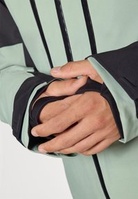 Jachetă verde deschis și neagră cu țesătură texturată, având manșete elastice, buzunare cu fermoar și un design elegant. Mâinile sunt poziționate aproape de manșete.