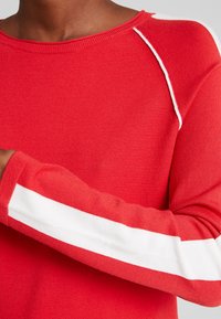 Maglione rosso con colletto a costine, caratterizzato da una striscia bianca lungo la manica e cuciture a contrasto sulla spalla. Texture liscia.