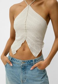 Kobieta ubrana w kremowy, fakturowany top na halter z falbaniastymi brzegami, połączony z jasnoniebieskimi dżinsami, z rękami w kieszeniach.