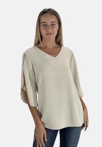 Top beige a V con maniche corte e morbide, realizzato in un tessuto texturizzato. L'orlo è leggermente curvato, abbinato a jeans in denim scuro.