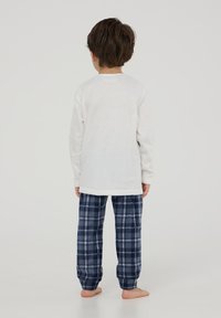 Biała bluzka z długim rękawem o miękkiej teksturze, zestawiona z granatowymi, kraciastymi spodniami do piżamy z niebieskimi i białymi kratkami. Dziecko stoi boso, widok z tyłu.