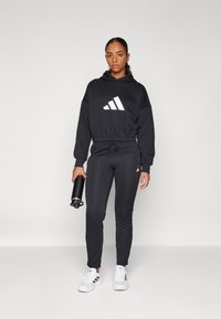 Sweatshirt preto com o logo branco da Adidas, combinado com calças de jogging pretas. O modelo segura uma garrafa de água preta, usando ténis brancos. Ajuste atlético padrão.