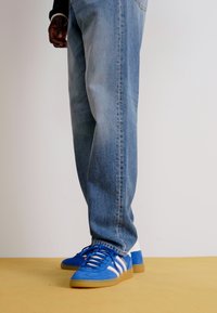 Modre športne čevlje z belimi črtami, kombinirani z svetlo modrimi jeans hlačami. Čevlji imajo teksturiran nubuk material in ravno gumijasto podplato.