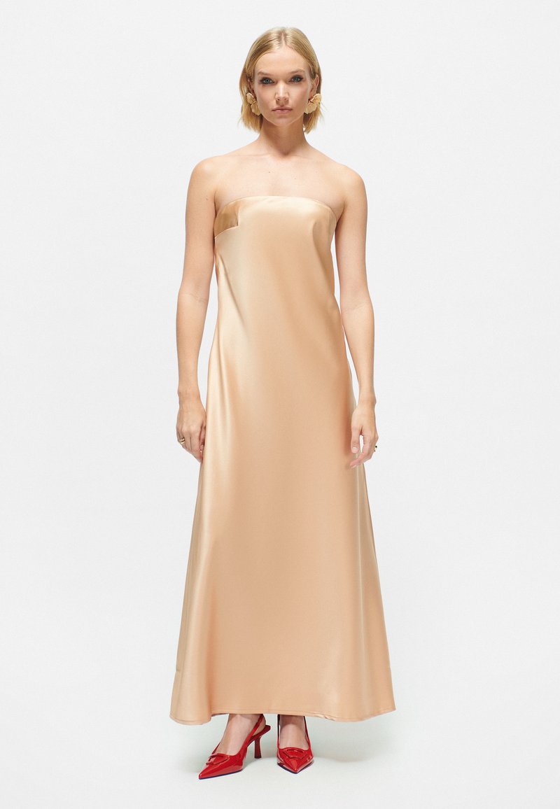 Halterlös satin klänning i mjuk beige, golvlång med en smidig silhuett. Kombinerad med glansiga röda högklackade skor för kontrast.