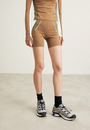 Personne portant un cuissard de vélo marron et olive à blocs de couleurs, un haut à manches longues assorti, des chaussettes noires et des chaussures de running grises sur un sol uni.