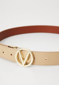 Bežs ādas josta ar apaļu zelta sprādzi, kurai ir "V" dizains. Gluda tekstūra ar vairākiem caurumiem regulēšanai.