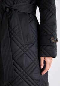 Μαύρο καπιτονέ παλτό με ζώνη στη μέση, διαθέτοντας διαγώνια ραφή, κουμπωτό μανίκι και ετικέτα λογότυπου στο πλάι. Λειαφή υφή.