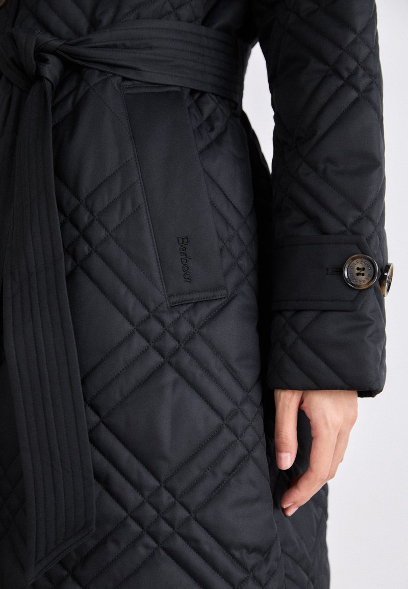 Cappotto nero trapuntato con vita regolabile, caratterizzato da cuciture diagonali, polsini con bottone e un'etichetta con logo sul lato. Tessuto liscio.