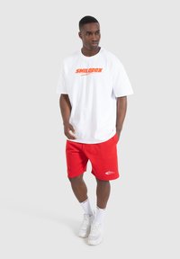 Witte T-shirt met korte mouwen en een rood "SMILODON" logo, gecombineerd met rode shorts met een wit logo. Model draagt witte sneakers en sokken.