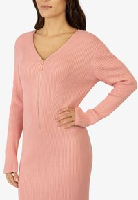 Femme portant une robe rose à manches longues côtelées, avec une fermeture éclair dorée à l'avant et un décolleté en V, main posée sur la hanche, fond neutre.