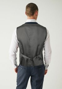 Gilet en satin gris avec un ajusteur arrière, porté sur une chemise blanche, associé à un pantalon bleu à motifs, avec deux poches arrière.