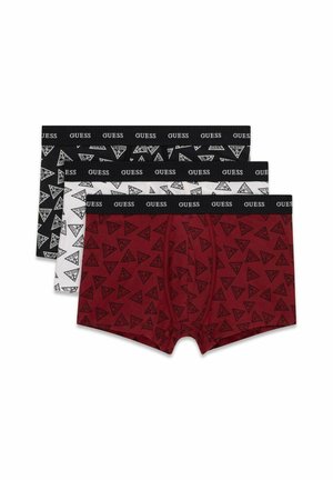 Homme musclé portant un boxer rouge avec des logos et motifs Guess noirs, ajustant la ceinture, debout devant un fond gris uni.
