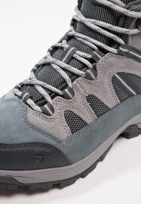 Stivale da hiking grigio caratterizzato da una combinazione di materiale scamosciato e mesh. Sistema di allacciatura dettagliato con rinforzi e suola testurizzata.