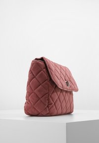 Borsa a mano in velluto mauve trapuntato con chiusura a patta e fibbia argentata, appoggiata su una superficie bianca contro uno sfondo leggero e semplice.