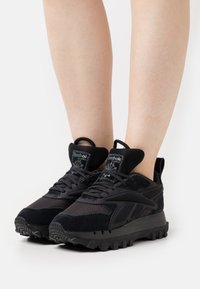 Chaussures de sport noires en mesh et suède ; caractéristiques : semelle texturée, soutien de la cheville rembourré, et un logo sur la languette.