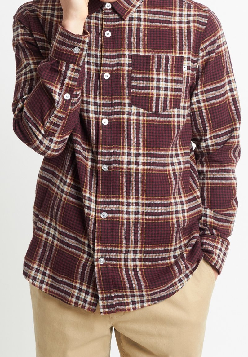 Homme portant une chemise à carreaux bordeaux, beige et blanche avec un pantalon beige, main dans la poche, sur fond blanc.