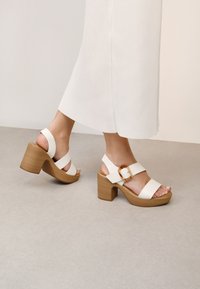 Sandalias de tacón alto blancas con correas anchas, un tacón de bloque de madera y un acento de hebilla dorada. Textura suave y diseño abierto que revela los dedos de los pies.