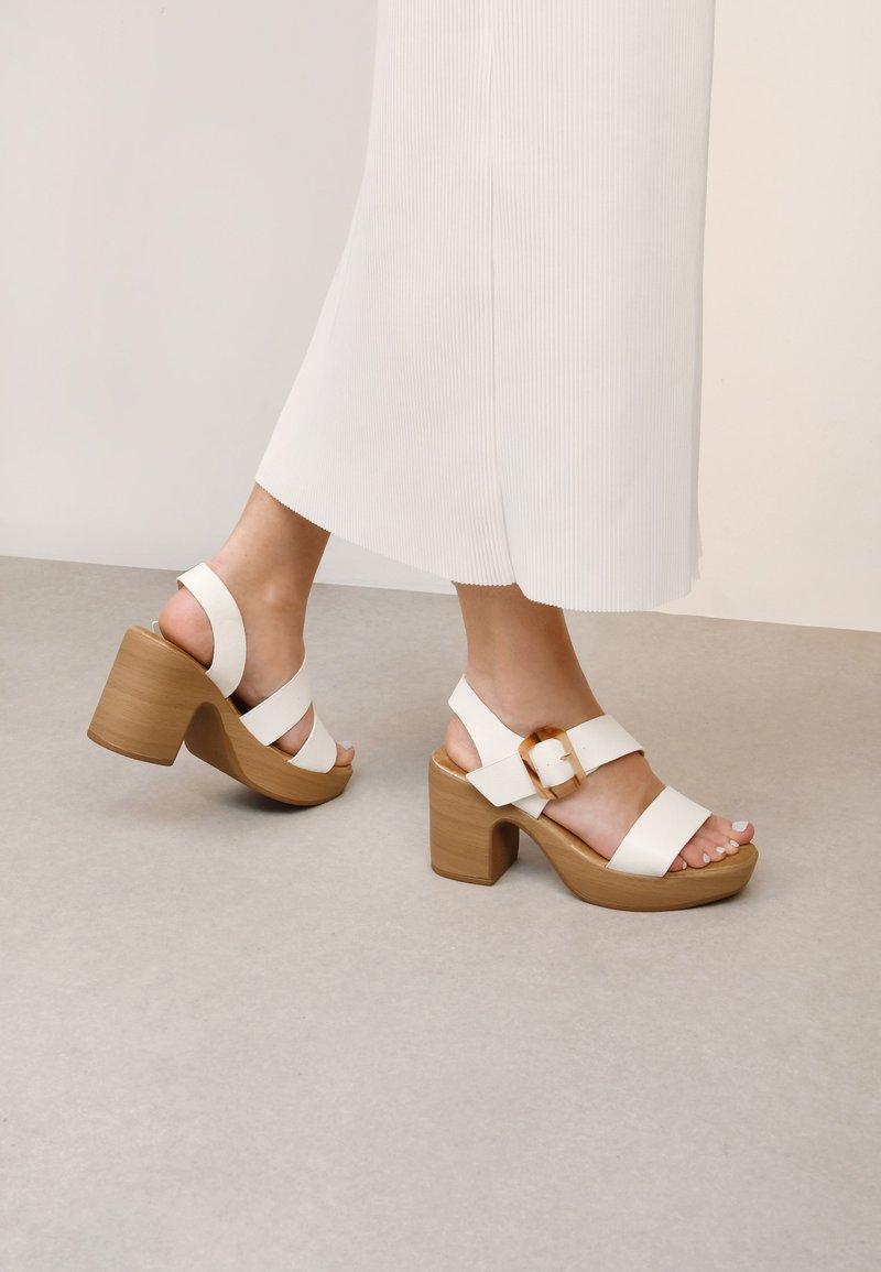 Sandalias de tacón alto blancas con correas anchas, un tacón de bloque de madera y un acento de hebilla dorada. Textura suave y diseño abierto que revela los dedos de los pies.