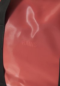 Punainen vedenpitävä materiaali kiiltävällä pinnalla, jossa on kohokuvioitu "RAINS" -logo. Mustat hihnat sivuilla tarjoavat rakenteellista tukea.