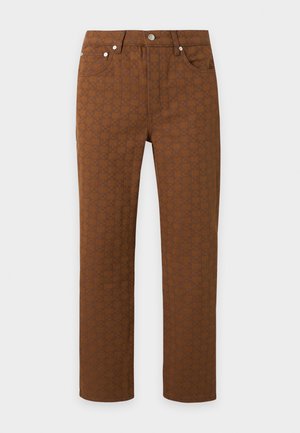 Jeune homme debout portant un polo noir, un pantalon marron à motifs et des mocassins marron foncé sur un fond gris uni.