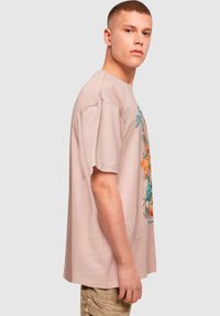 Světle růžové oversized tričko vyrobené z bavlny, s grafickým květinovým potiskem v modré a oranžové na přední straně. Krátké rukávy, kulatý výstřih.