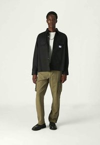 Modelo masculino vestindo uma camisa preta com botões sobre uma camisa branca, calças cargo verde-oliva, sapatos pretos e um colar de corrente prateado, em um fundo claro e simples.
