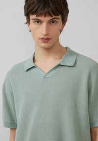 Polo shirt tricoté à manches courtes de couleur vert clair avec un motif texturé, présentant un col en V et un col contrastant dans une teinte légèrement plus claire.