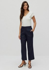 Granatowe spodnie o kroju dopasowanym z bocznymi kieszeniami, wysokim stanem i luźnym fasonem, w połączeniu z białą bluzką z krótkim rękawem i dekoltem w serek oraz czarnymi sandałami na obcasie.