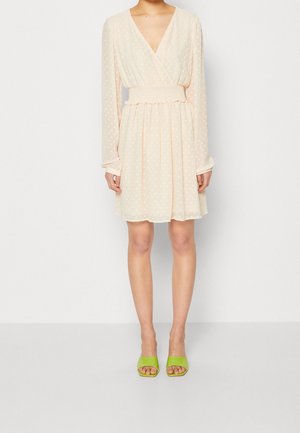 Kvinde iført en knælængde, langærmet, cremefarvet kjole med subtile polkaprikker og grønne åbne hælede sandaler.