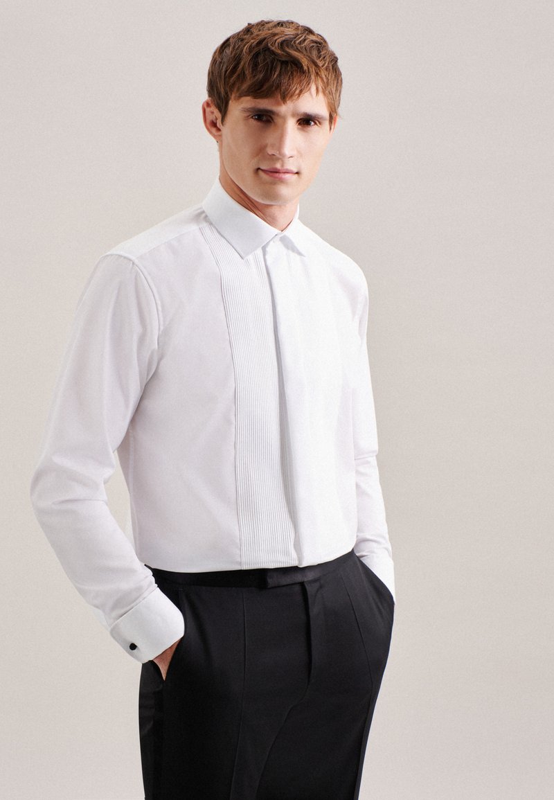 Young man wearing a white pleated tuxedo shirt and black pants with hands in pockets against a plain background.