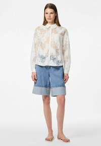 Kant blouse met bloemenpatronen in perzik- en blauwtinten, gecombineerd met denim culottes met een rauwe zoom en contrasterende stiksels. Ontblote sandalen.