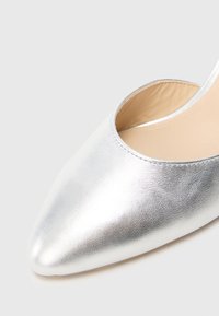 Sapato metálico prateado com bico pontiagudo e acabamento em couro liso, apresentando um lado aberto e uma palmilha bege, destacando um design elegante e moderno.