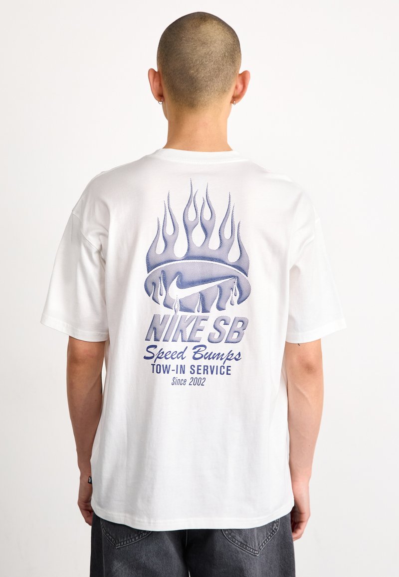 Bílý bavlněný tričko s modrým grafickým designem na zadní straně, včetně plamenů a textu "NIKE SB Speed Bumps". Krátké rukávy, volný střih.