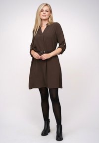 Braunes Kleid mit V-Ausschnitt, aufgerollten Ärmeln und einem schwarzen Gürtel. Getragen mit schwarzen Knöchelstiefeln und schwarzen Strumpfhosen vor einem neutralen Hintergrund.