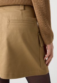 Gros plan sur une personne portant un pull marron en tricot et une jupe beige avec une poche fendue à l'arrière et une poche à rabat sur le côté.