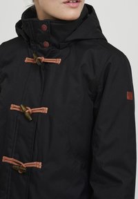 Schwarze Jacke mit hohem Kragen, ausgestattet mit braunen Seilknöpfen und Bambusakzenten. Glatter Stoff mit leicht strukturierter Oberfläche.