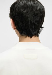 Weißes Baumwollhemd mit tiefem Kragen und einem rechteckigen Etikett auf der Rückseite. Der Stoff wirkt glatt mit einer subtilen Textur.