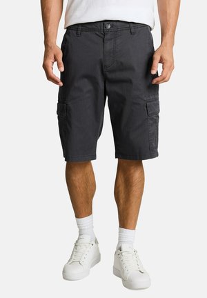Mann trägt schwarze Cargoshorts, weiße Socken und weiße Turnschuhe, steht mit entspannten Händen an den Seiten.